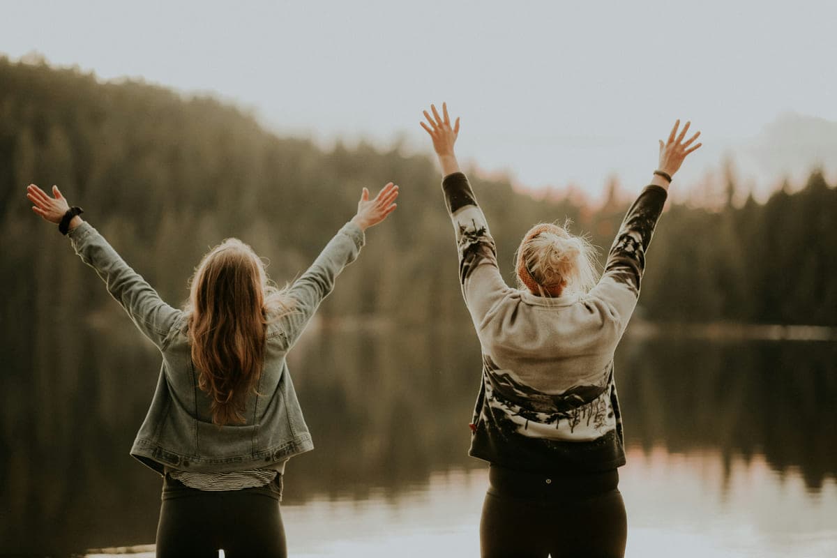 Two people seen from behind with their arms raised, facing a lake and forest.