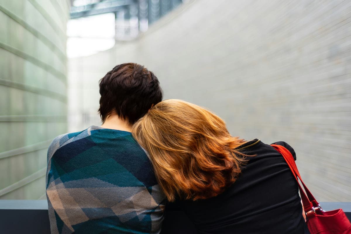 Two people sitting close together seen from behind, one resting their head on the other's shoulder.