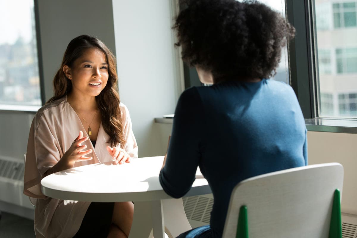 Two people sitting at a small round table by a window, one facing the camera mid-conversation.
