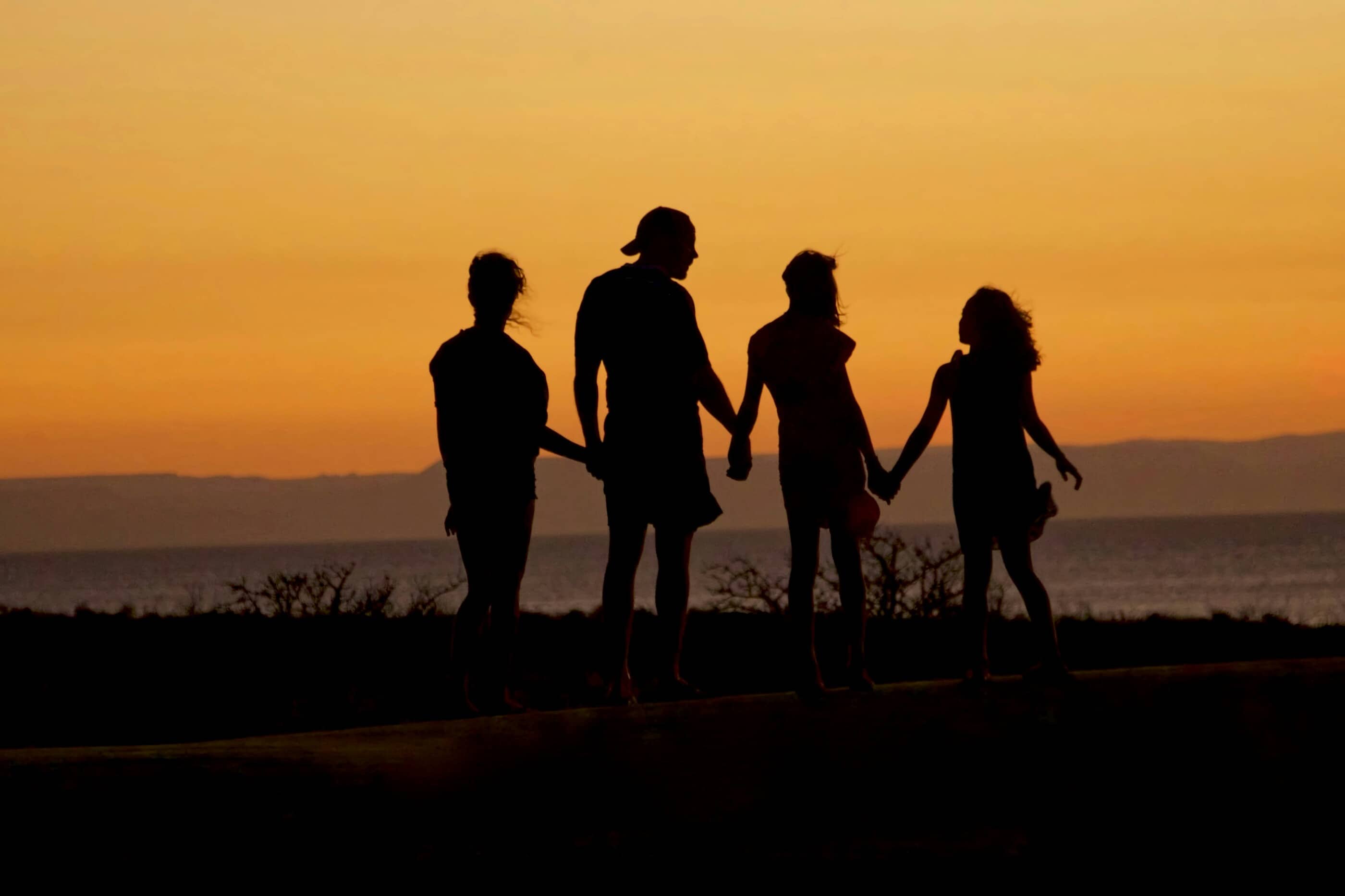 A family of four holding hands, silhouetted against an orange sunset by the water.