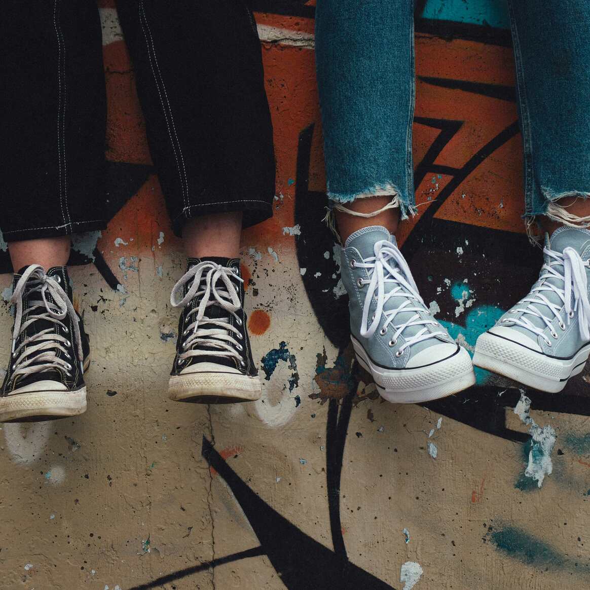 Two pairs of legs in high-top trainers, dangling in front of a graffitied concrete wall.