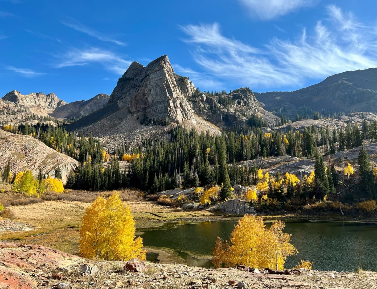 A mountain lake with golden aspen trees below a pointed granite peak under a blue sky.