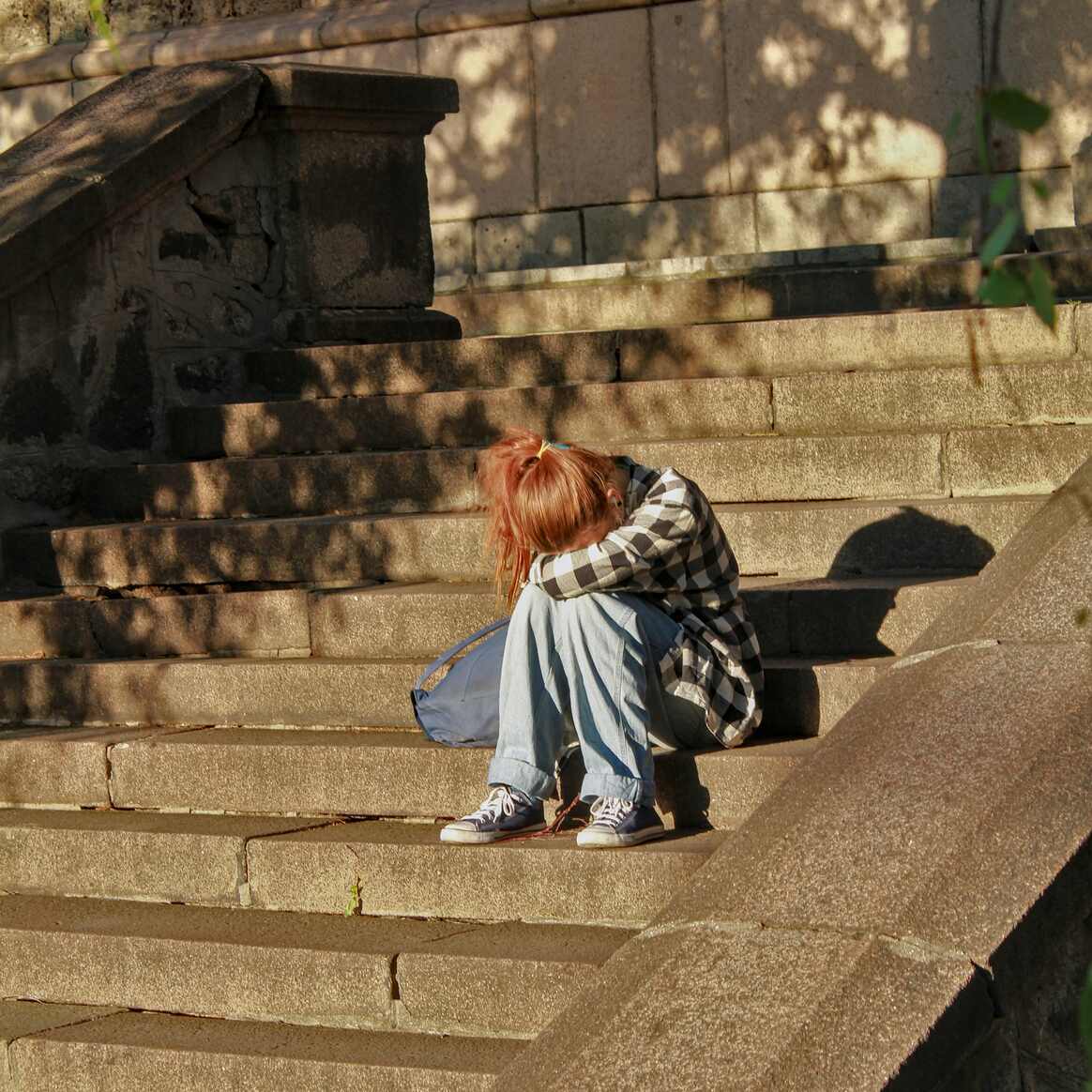 A person sitting on stone steps with their head resting on their folded arms, seen from behind.