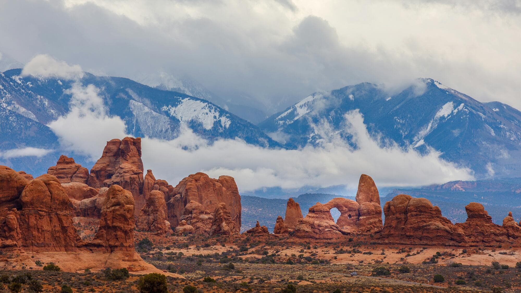 Red rock formations and a natural arch below snow-capped mountains and low cloud.