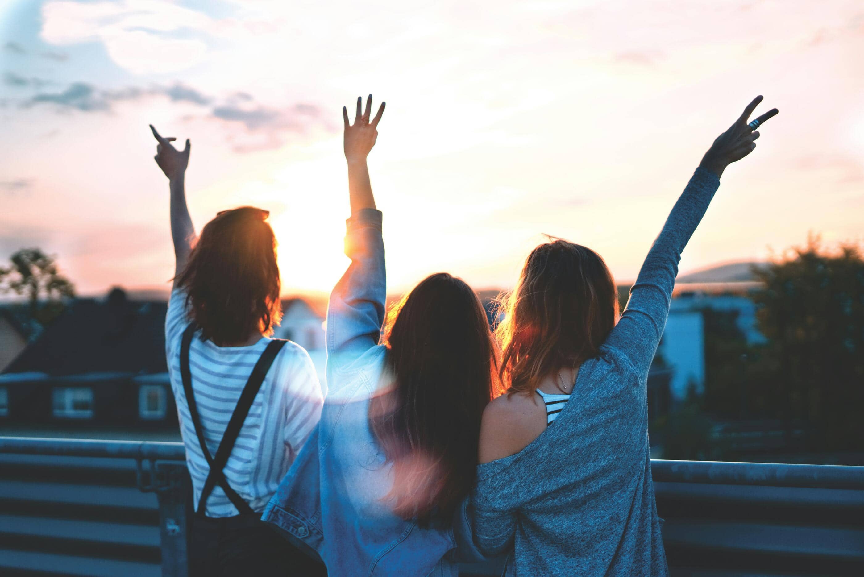 Three young people raising their arms toward a sunset over rooftops, seen from behind.
